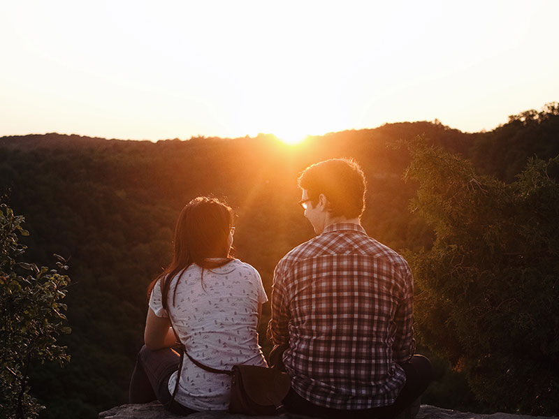A couple on a first date asking each other first date questions while sitting in front of a sunset.