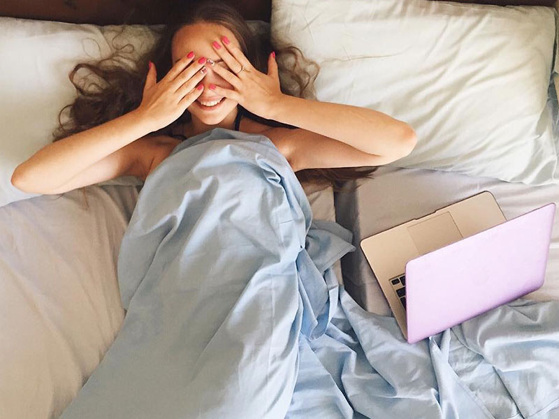 A woman suffering from online dating fatigue in bed covering her eyes next to her computer.