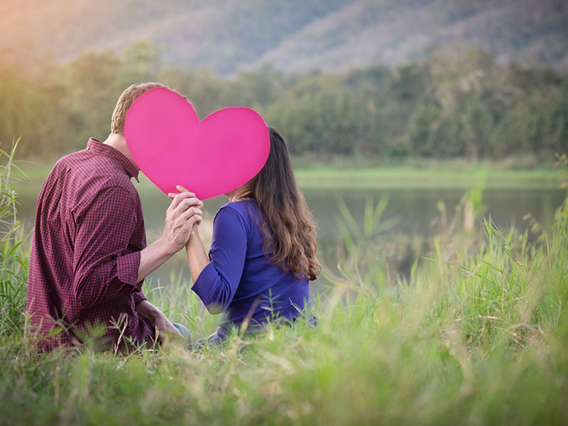A couple getting creative in how to show affection by kissing behind a pink heart in a field.