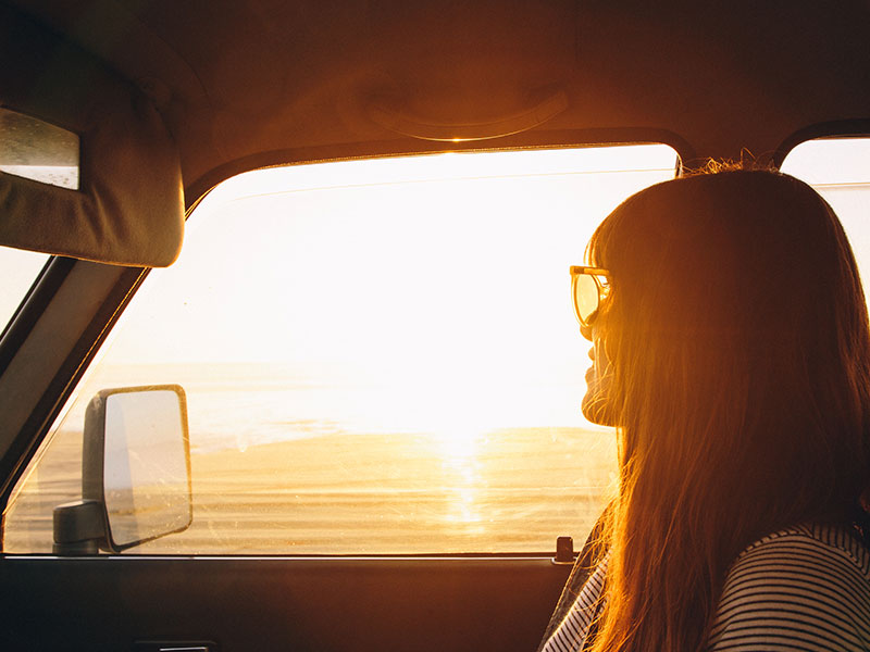 A women who is letting go of someone you love, in a car looking out the window at sunset on an open road.