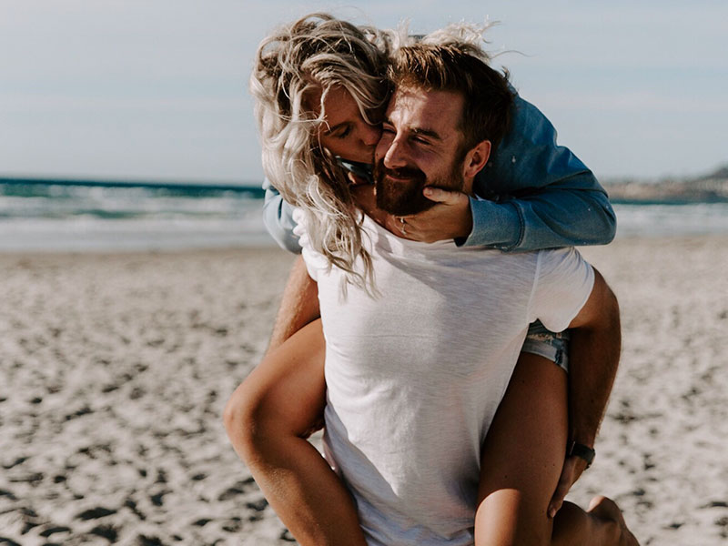 A woman who figured out what single men want, laughing and kissing her boyfriend’s cheek on the beach.
