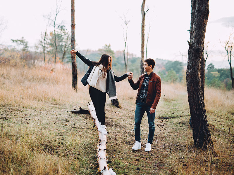 A girl who's wondering "is he thinking about me?" balancing on a log while the guy she likes helps her and looks on with a smile.