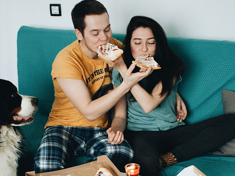 A guy who learned how to flirt with a girl, feeding a girl pizza as she laughs and takes a bite.