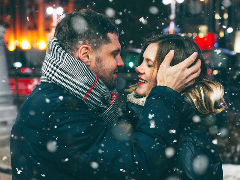 A guy experiencing blind love, kissing his date in the snow as they both smile at each other.