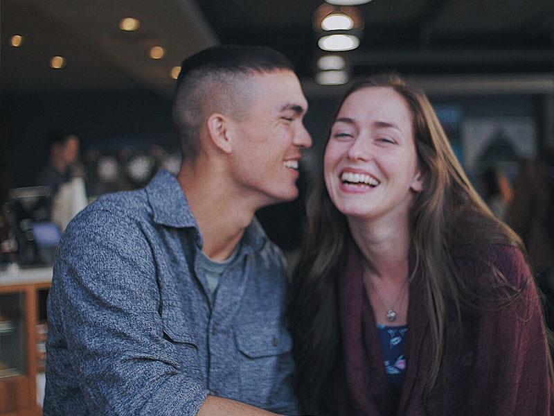 A guy who learned how to talk to woman, whispering in her ear at a coffee shop as she laughs.