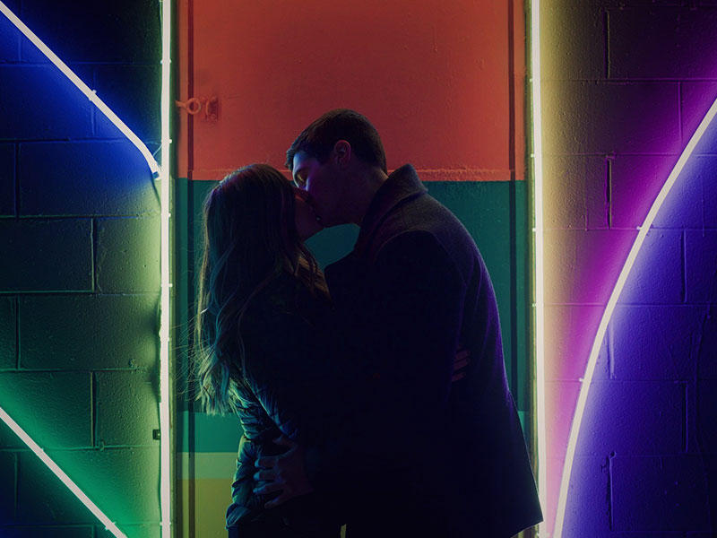 A women wondering "Am I in love?" as she dramatically kisses her boyfriend in front of a neon sign.