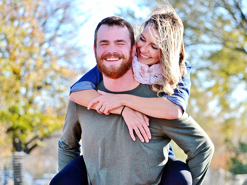 A man laughing as he carries his girlfriend on his shoulders as she looks at him and wonders, "how do men fall in love?"
