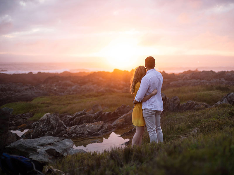 A woman wondering how men in love act, hugging her boyfriend while they watch the sunset.