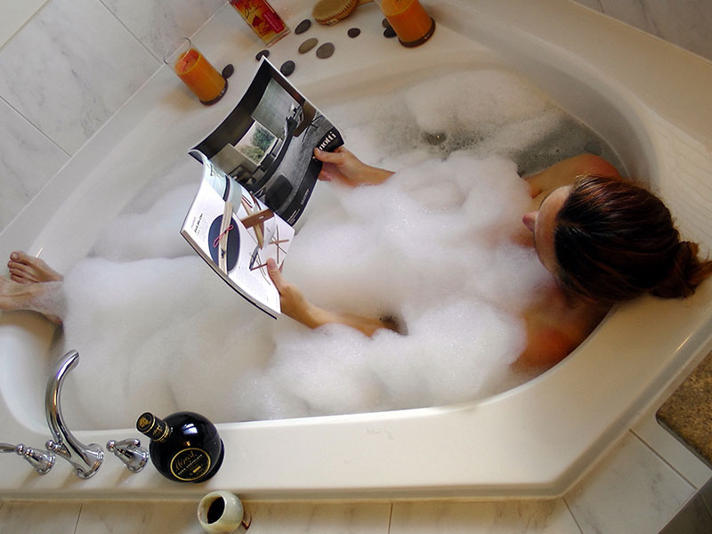 A woman in a bathtub reading a magazine in a bubble bath relaxing.