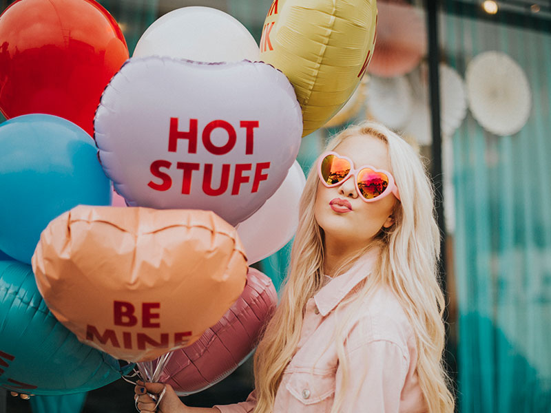 A girl with Valentine's Day balloons and heart sunglasses making a kissy face at the camera.