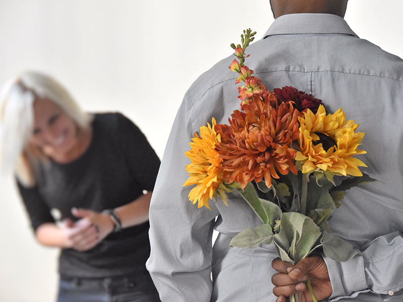A man dating after 50, holding flowers behind his back while his date laughs in front of him.