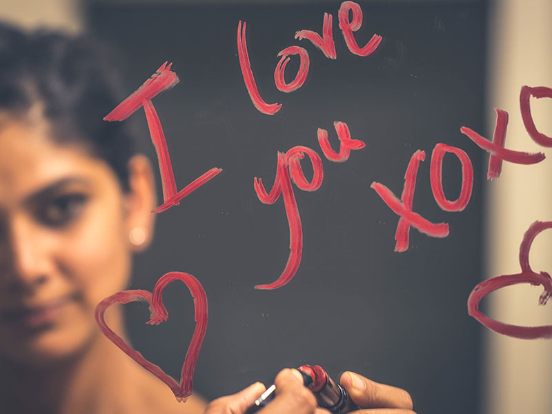 A woman writing, "I love you" on a mirror with lipstick, one of the romantic gestures for him that guys appreciate.