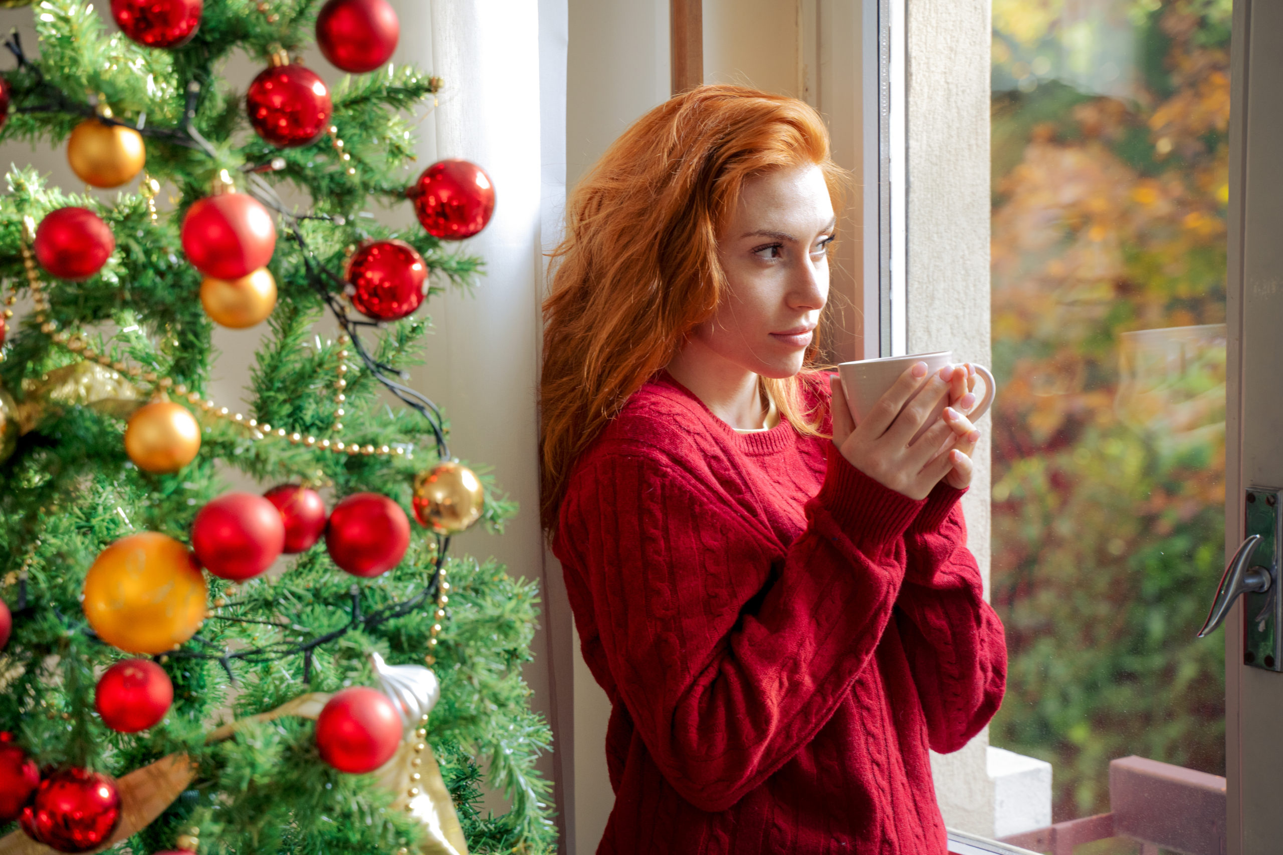 Pensive young woman standing by Christmas tree looking out of the window and thinking about how to survive the holidays after a breakup.