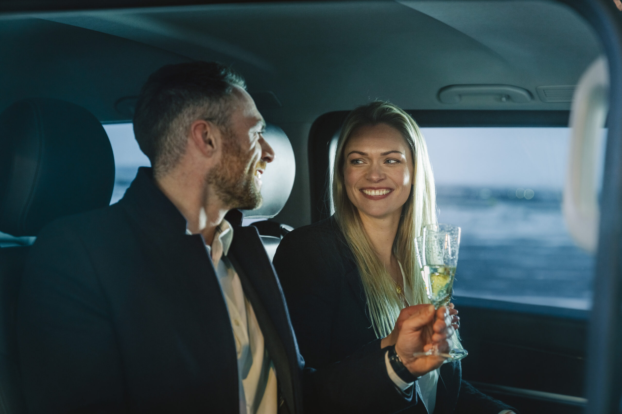 Young couple smiling at each other and experiencing millionaire dating while drinking wine and traveling in the back of a car.