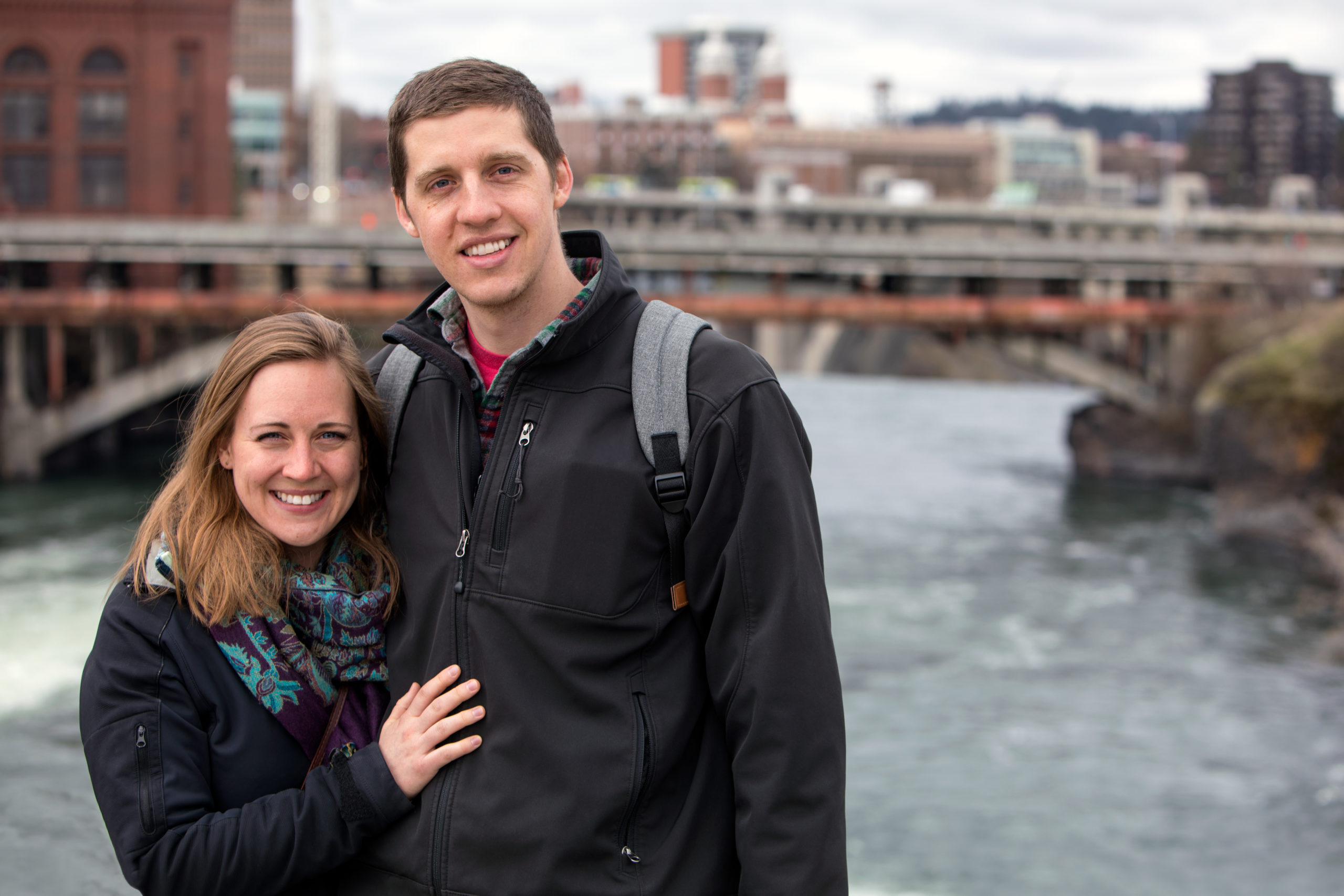Happy couple looking at camera and standing by a river while dating in Spokane.